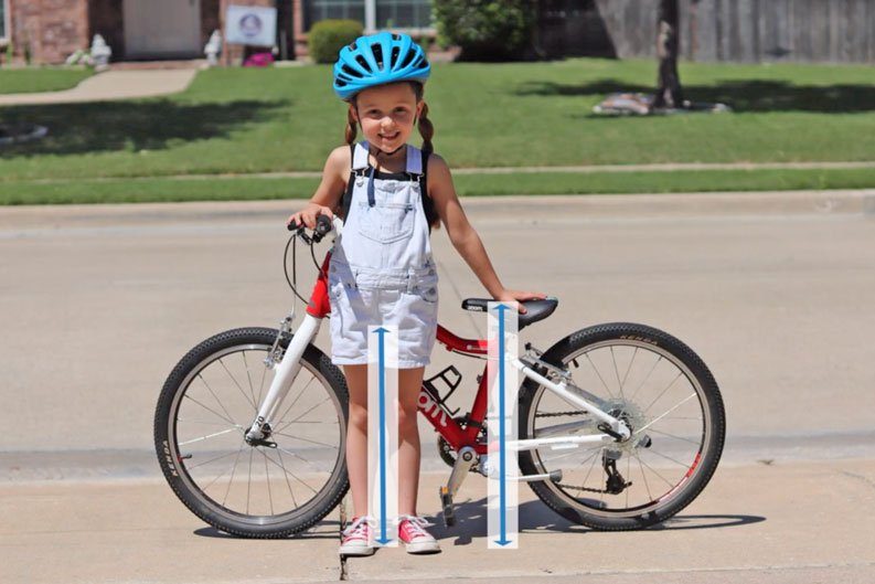 5 year old standing next to her bike. Arrows indicating inseam measurement and bike seat height measurement to help find correct kid bike size