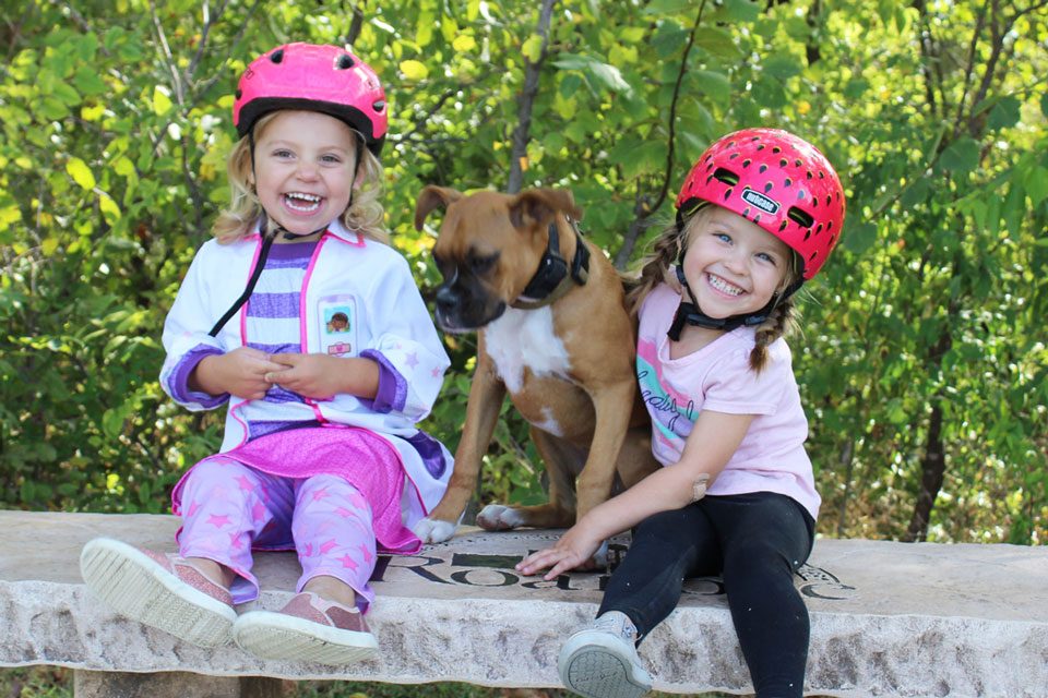 Two toddlers wearing bike helmets. One is wearing a pink Giro Scamp. The other is wearing a Strawberry Nutcase Baby Nutty.